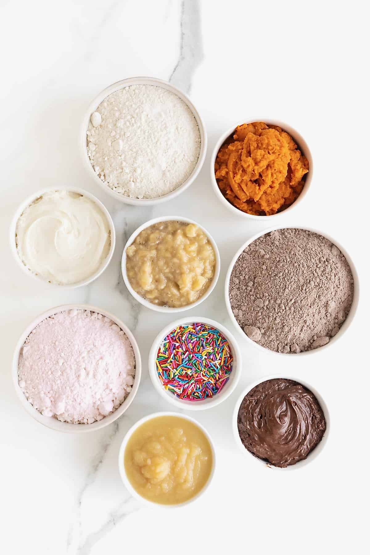 Ingredients to make cake mix donuts in small white dishes on a white marble counter.