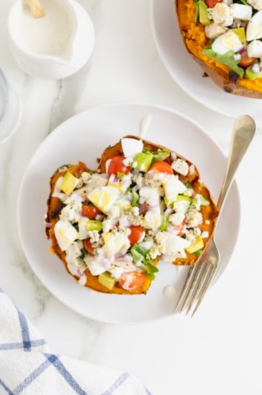 A sweet potato loaded with Cobb salad ingredients on a white dinner plate with a fork.