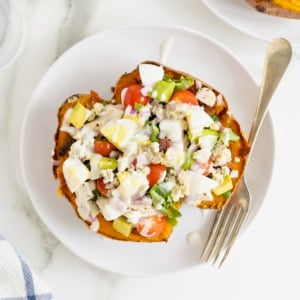 A sweet potato loaded with Cobb salad ingredients on a white dinner plate with a fork.