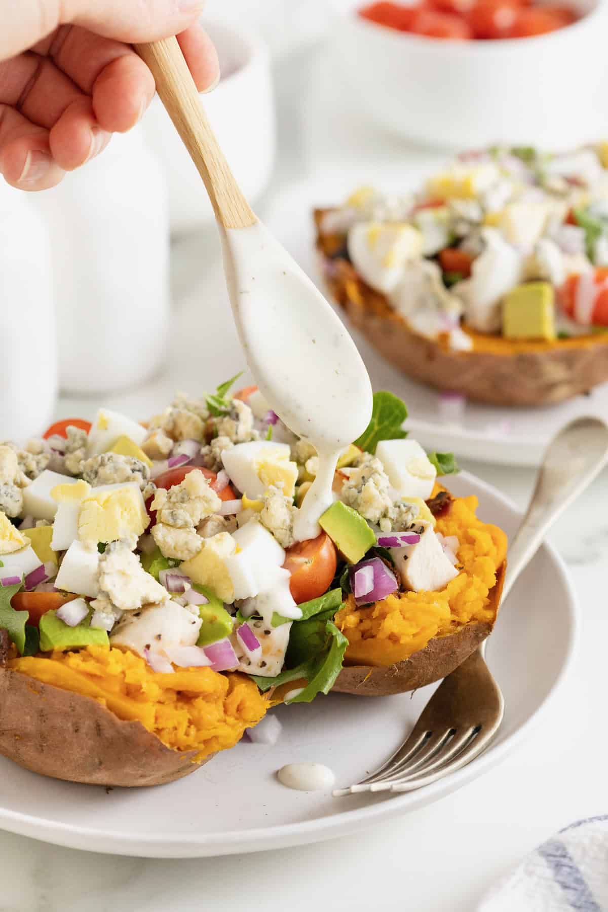 A Cobb salad sweet potato on a white dinner plate with a fork. Dressing is being drizzled onto the potato with a small wooden spoon.