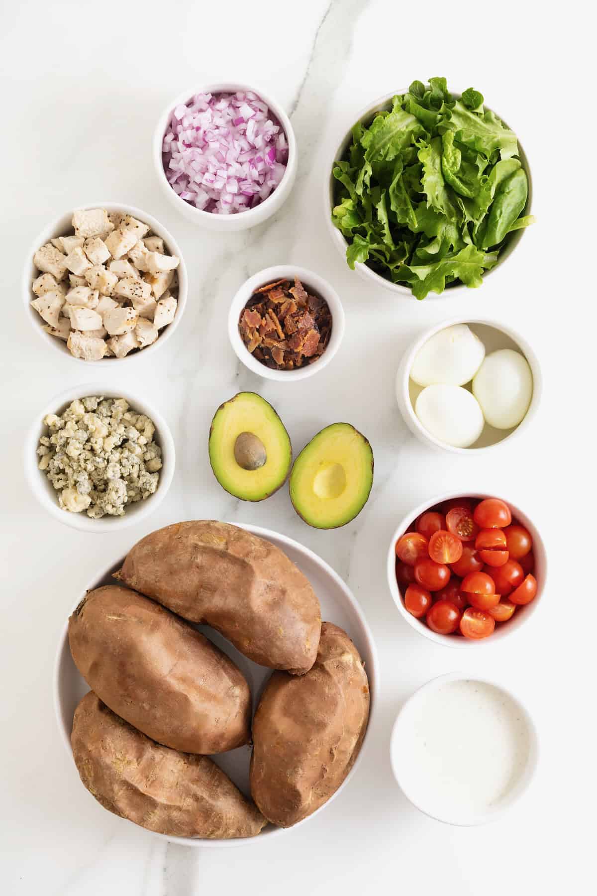 Ingredients to make a Cobb salad sweet potato in small white dishes on a white marble counter.