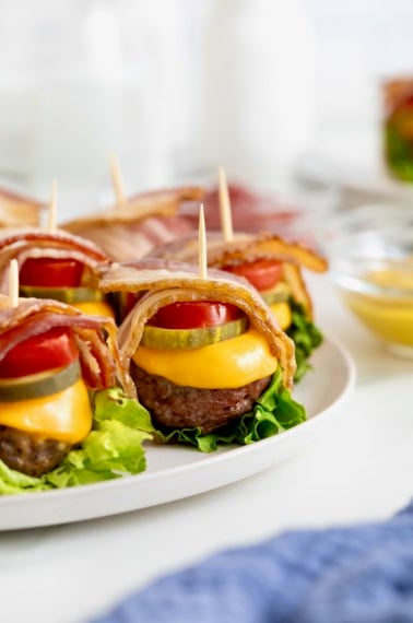 Six bacon wrapped cheese burger bites on a white platter.