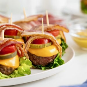 Six bacon wrapped cheese burger bites on a white platter.