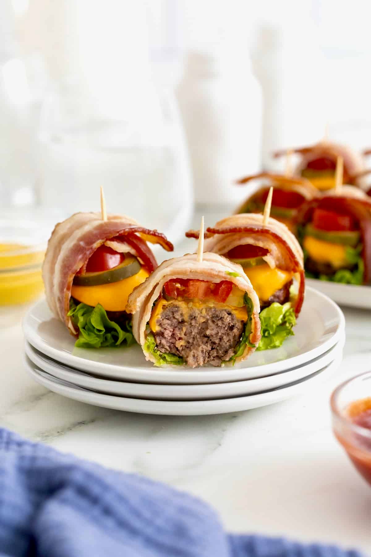 Three bacon wrapped cheeseburger bites on a stack of three white plates. The foremost burger has a bite out of it.