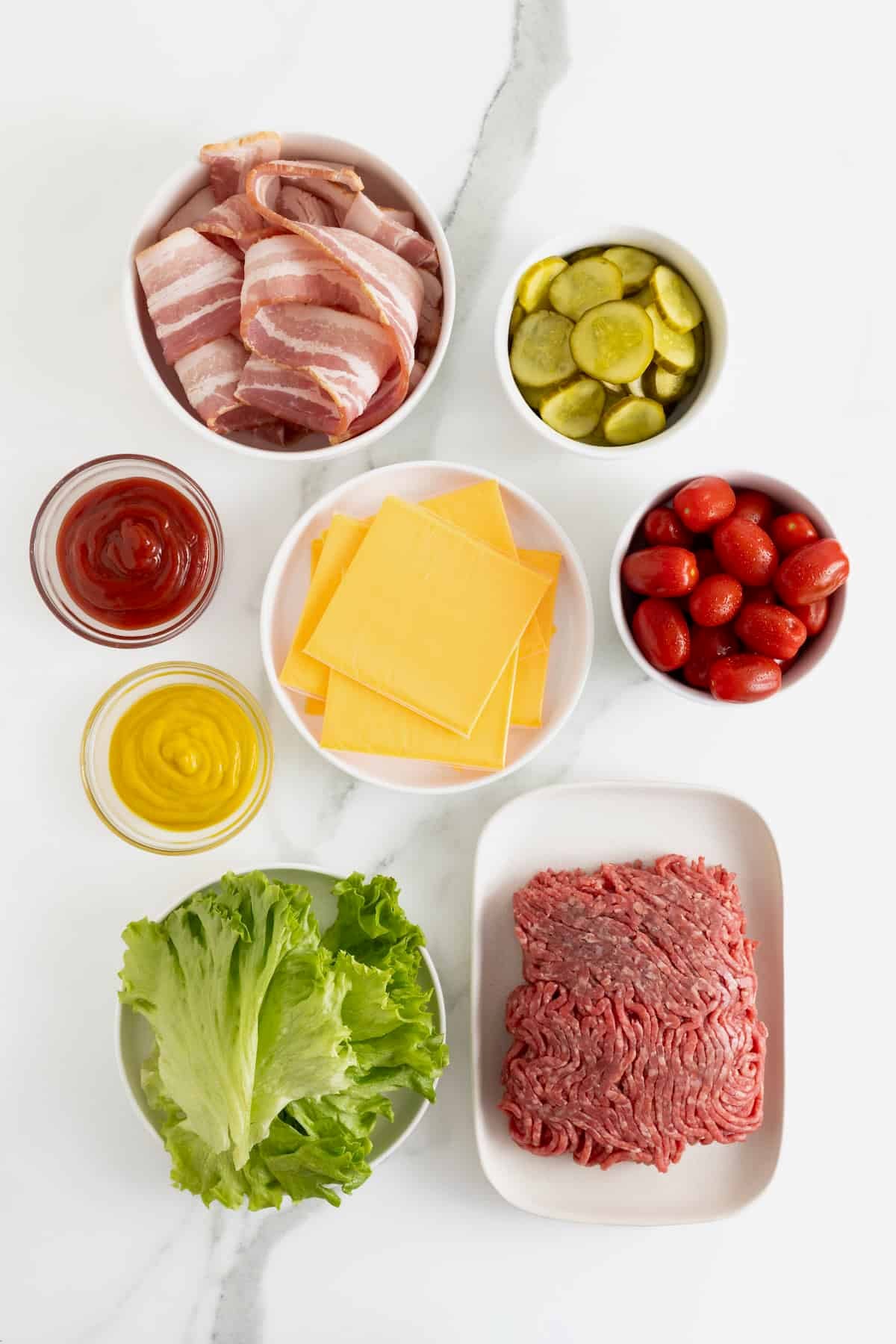 Ingredients to make bacon wrapped cheeseburger bites in small glass dishes on a white marble counter.