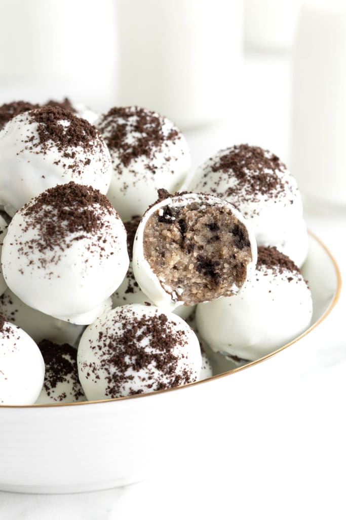 A large white serving bowl filled with cake balls coated in white coating and dusted with cookie crumbs. One of the cake balls has a bite out of it revealing the Oreo cookie cake inside.