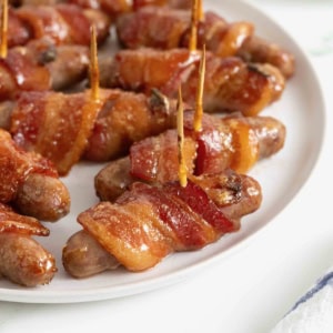 A white serving platter of breakfast sausages wrapped in bacon and secured with toothpicks.