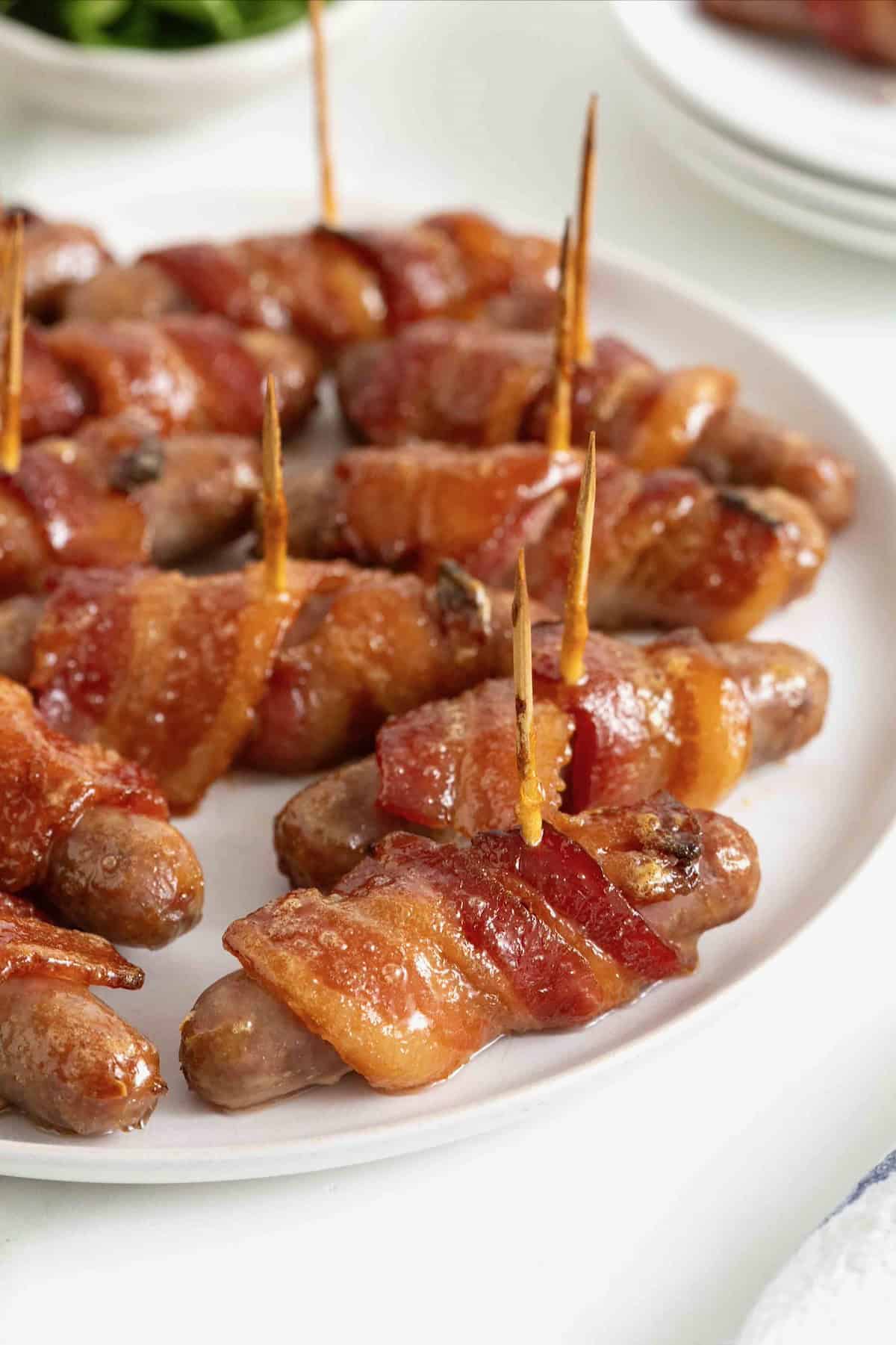 A white serving platter of breakfast sausages wrapped in bacon and secured with toothpicks.