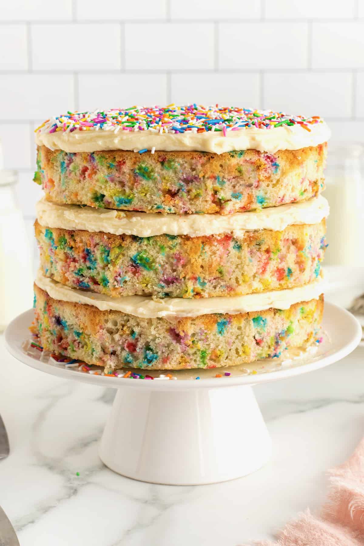 A white layer cake with rainbow sprinkles in the cake and white frosting on the top with more colorful sprinkles.