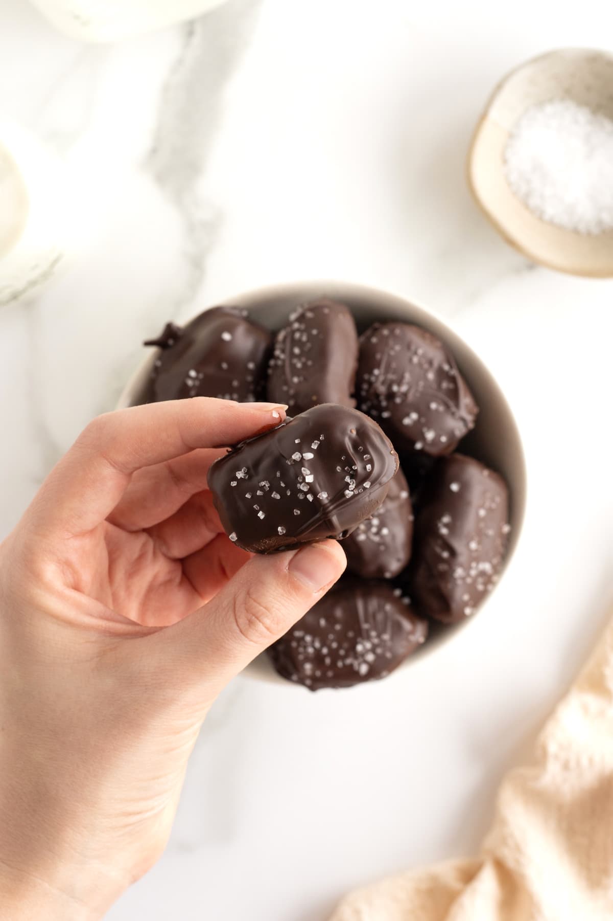 A bowl of chocolate covered dates sprinkled with sea salt and a hand holding one of the dates between two fingers. 