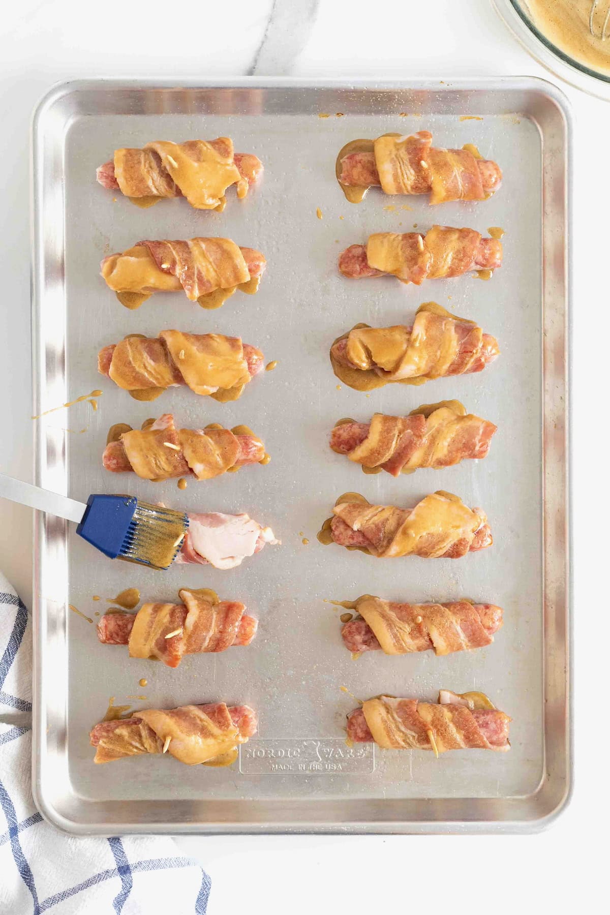 Bacon wrapped sausages being brushed with glaze on an aluminum baking sheet.