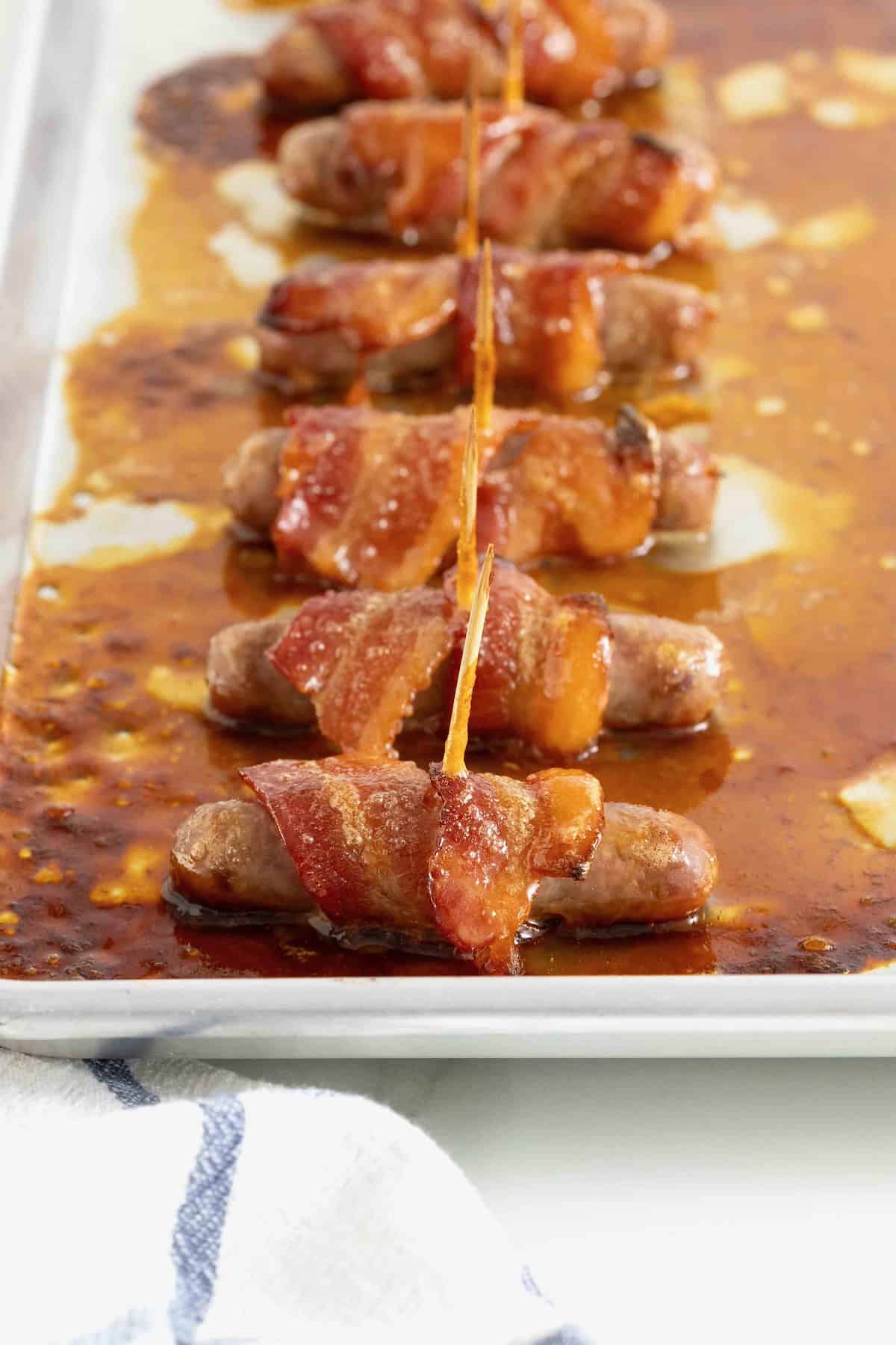 Bacon wrapped sausages on a rimmed aluminum  baking sheet.