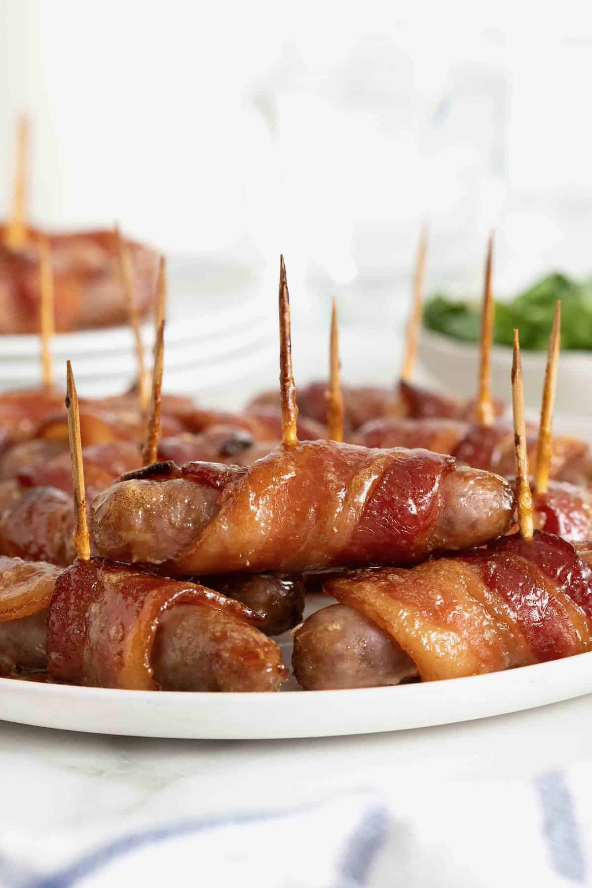 A white serving platter of breakfast sausages wrapped in bacon and secured with toothpicks.
