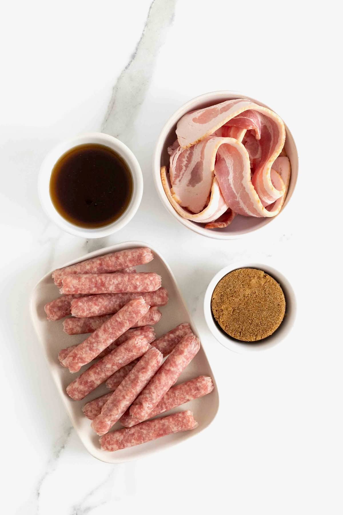 Ingredients to make maple glazed bacon wrapped sausages in small white dishes on a white marble counter.