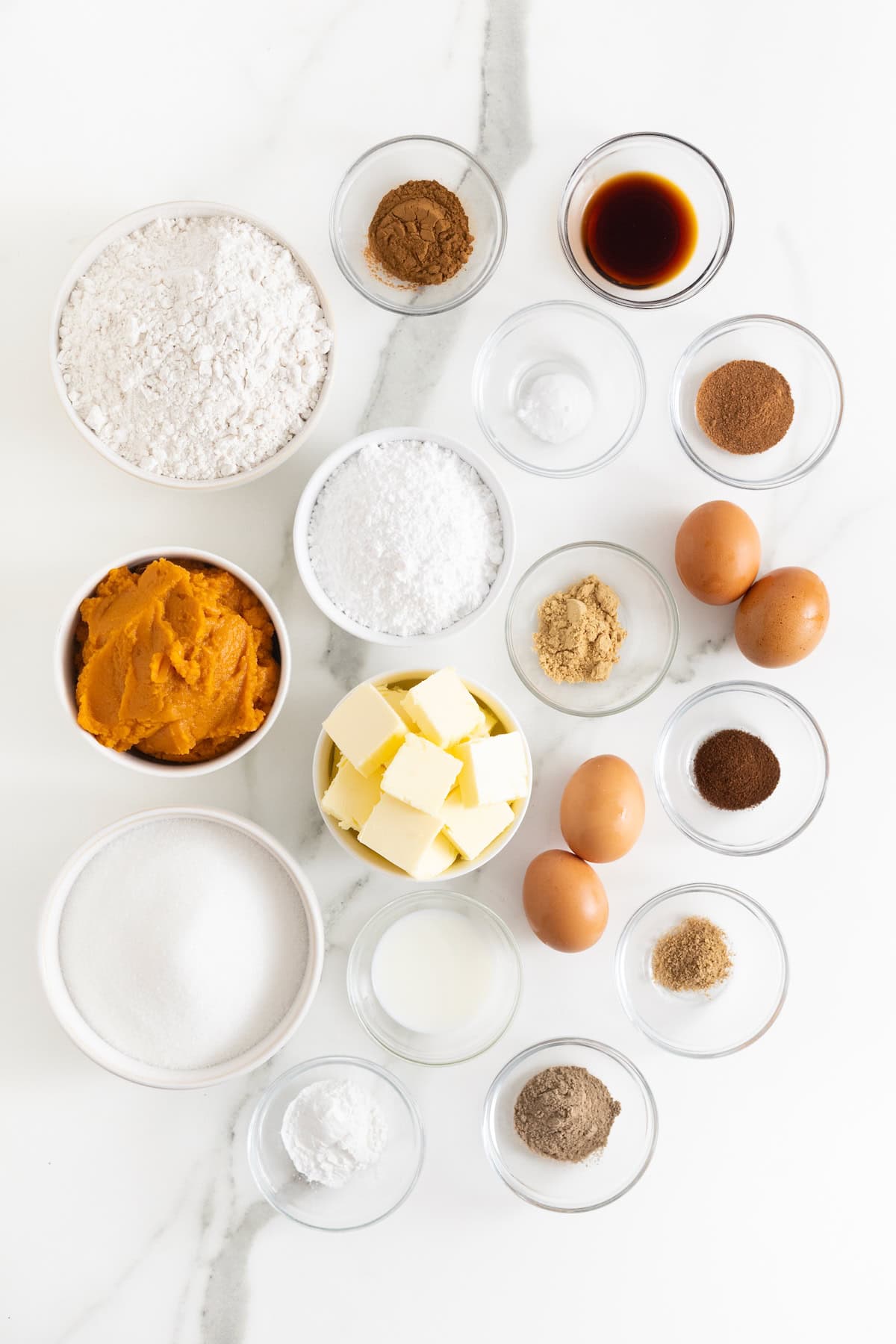 Ingredients to make a chai spiced pumpkin pound cake in small glass dishes on a white marble counter.