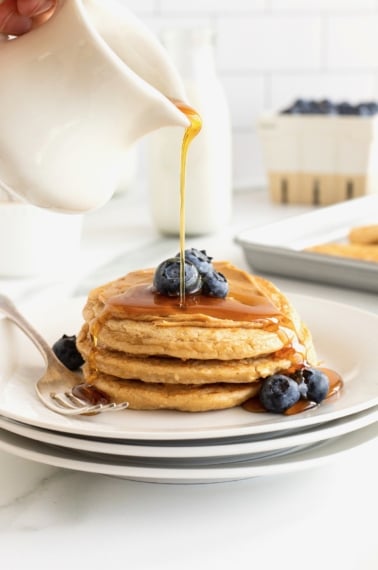 A stack of three protein pancakes smeared with peanut butter and topped with fresh blueberries and maple syrup.