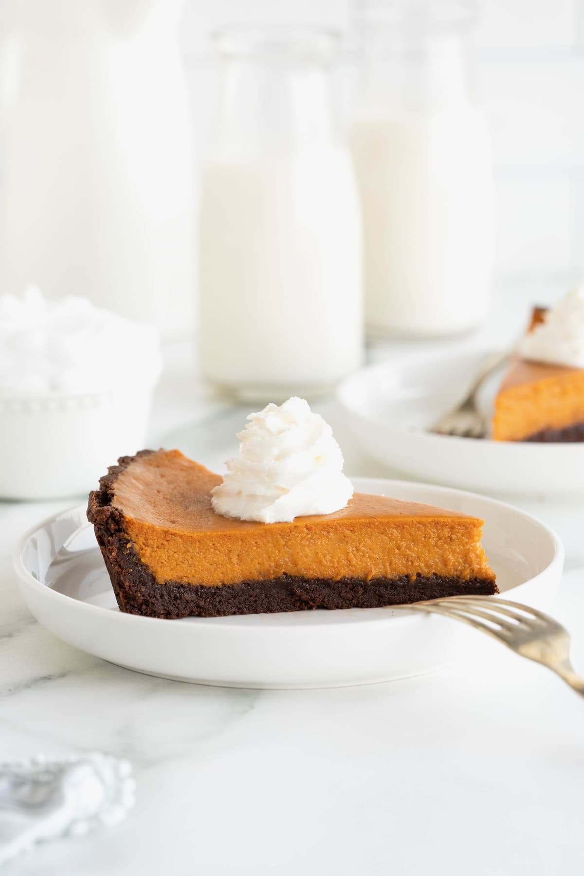 A slice of pumpkin tart with a chocolate crust on a white rimmed plate. The tart is topped with whipped cream.