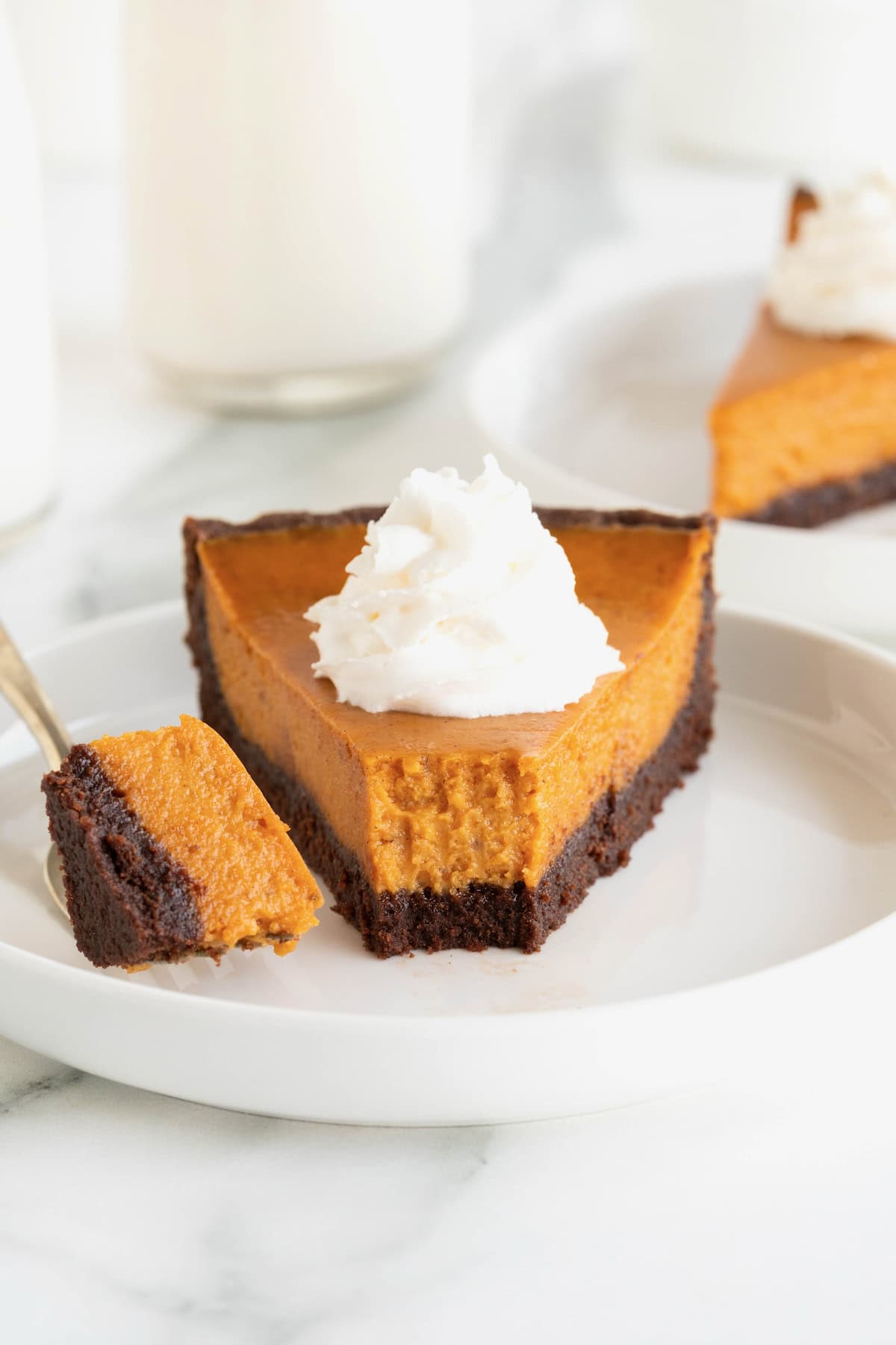 A slice of pumpkin tart with a chocolate crust on a white rimmed plate. The tart is topped with shipped cream.