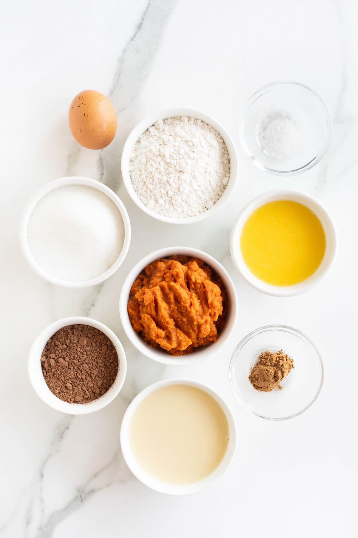 Ingredients to make dark chocolate pumpkin tart in small white glass dishes on a white marble counter.