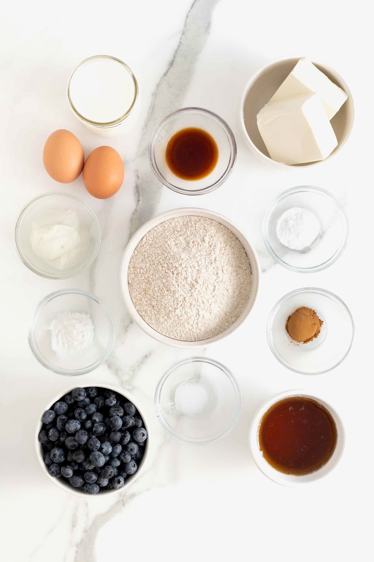 Ingredients to make whole wheat blueberry breakfast cupcakes in small glass dishes on a white marble counter.
