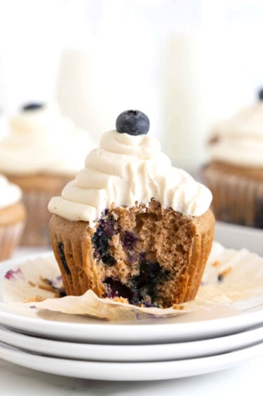 A blueberry cupcake with white cream cheese icing unwrapped on a white plate.