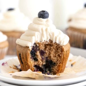 A blueberry cupcake with white cream cheese icing unwrapped on a white plate.