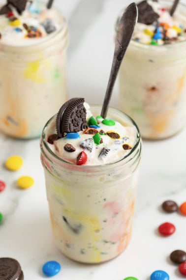 A homemade M&M Blizzard garnished with a piece of Oreo in a wide mouth jar. There is a spoon sticking out of the jar.