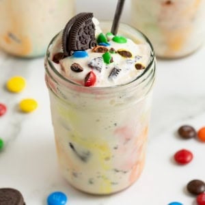 A homemade M&M Blizzard garnished with a piece of Oreo in a wide mouth jar. There is a spoon sticking out of the jar.