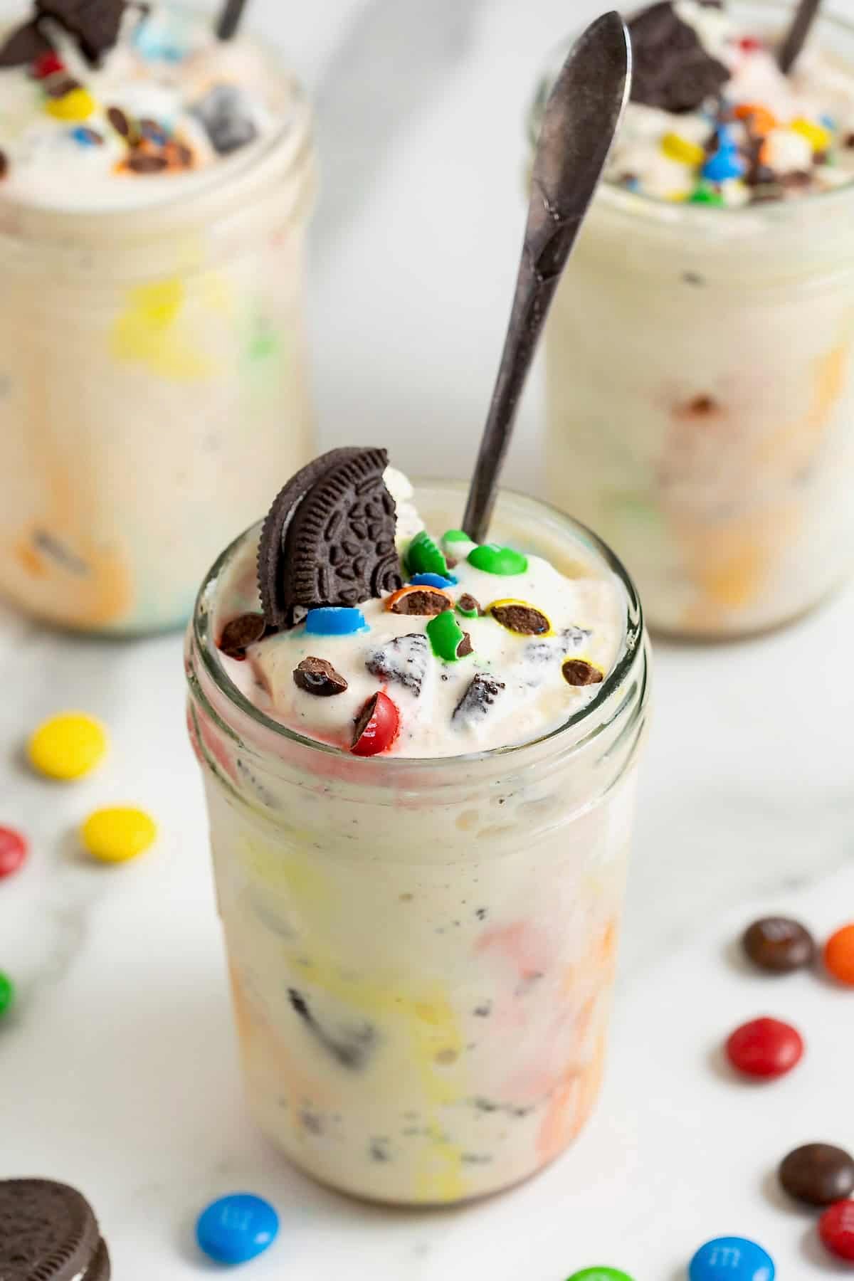 A homemade M&M Blizzard garnished with a piece of Oreo in a wide mouth jar. There is a spoon sticking out of the jar.