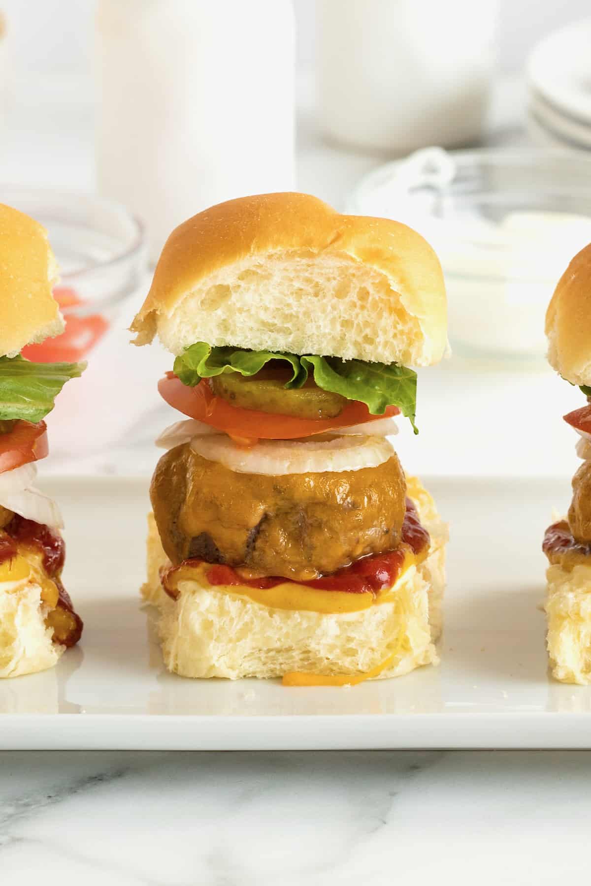 Three mini cheeseburgers with onion, tomato, pickle and lettuce on a white serving plate.
