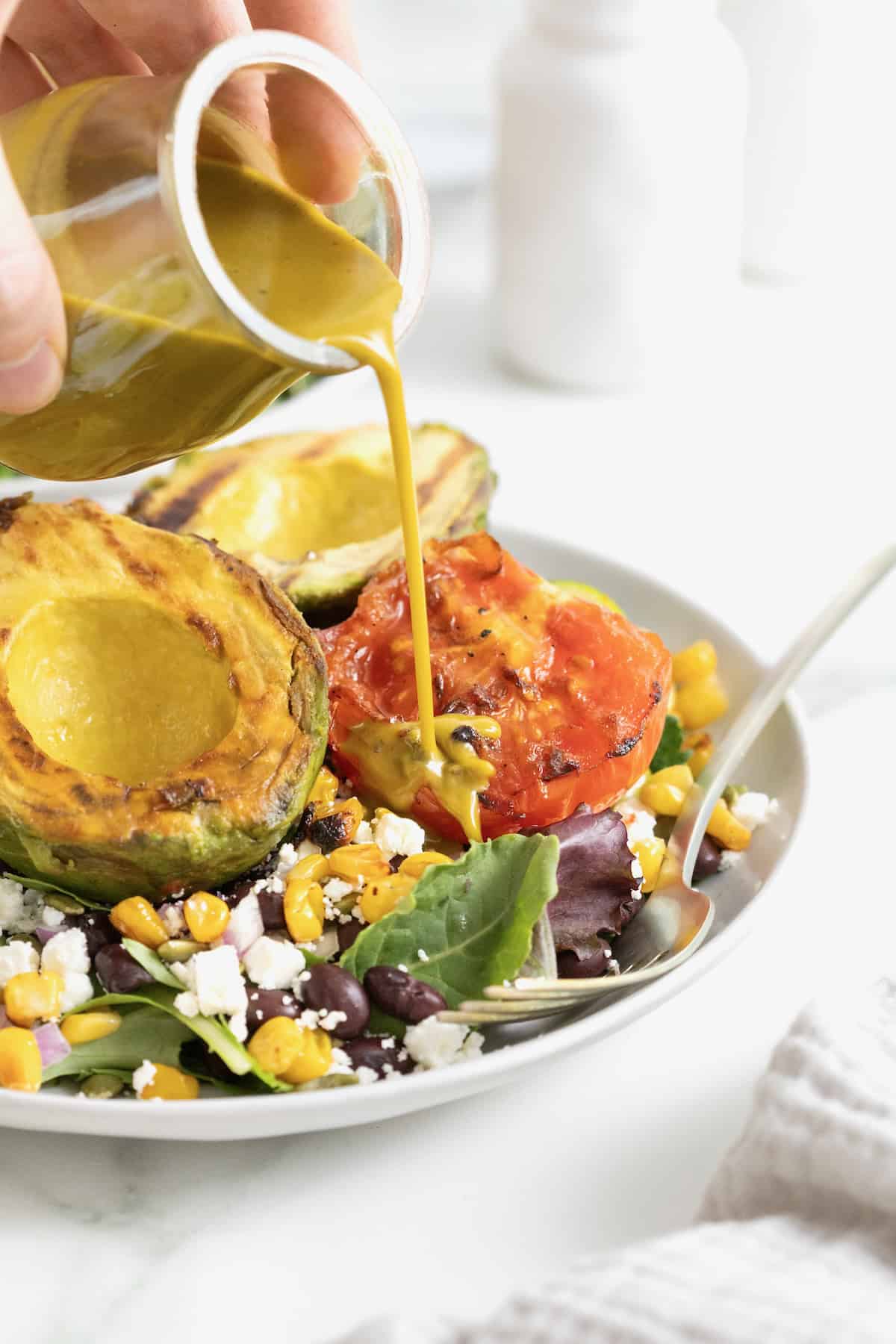 A chipotle salad dressing being poured over a grilled avocado and tomato salad.