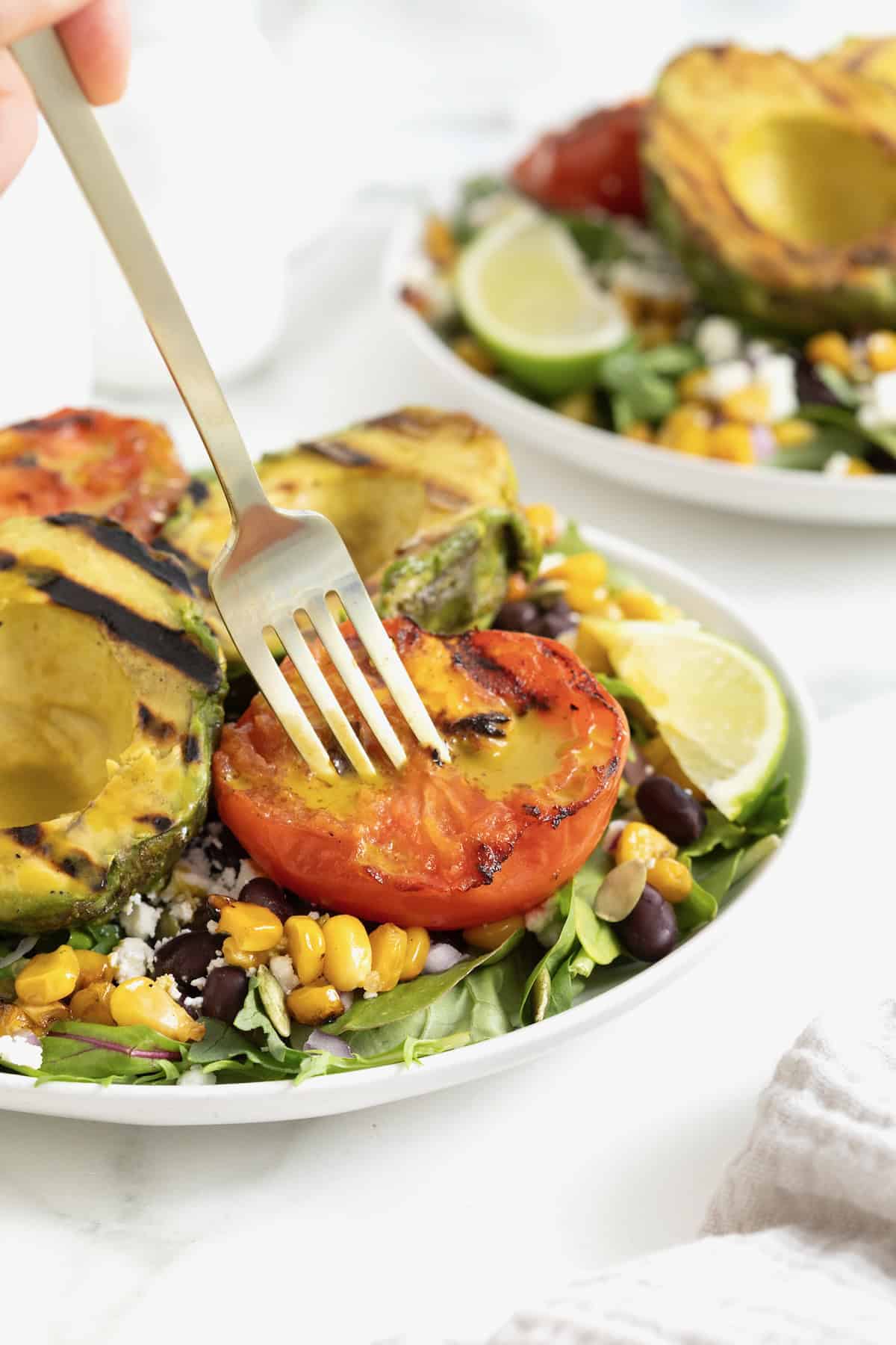 A fork piercing a grilled tomato half on a plate of salad.