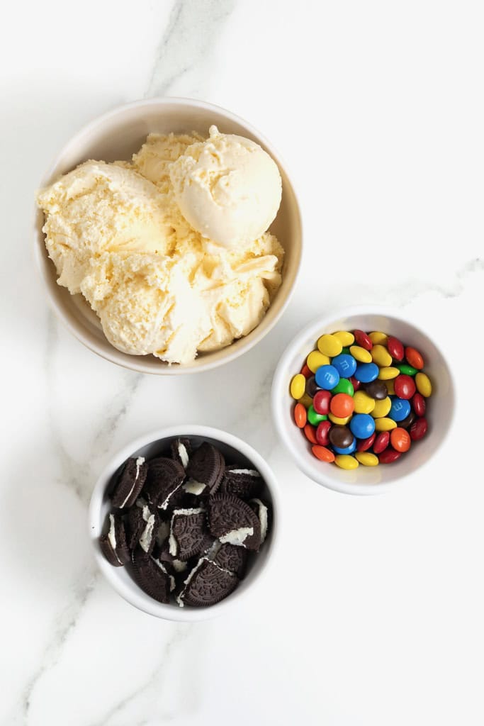 Ingredients to make a homemade Oreo and mini M&M blizzard in white dishes on a white marble counter.