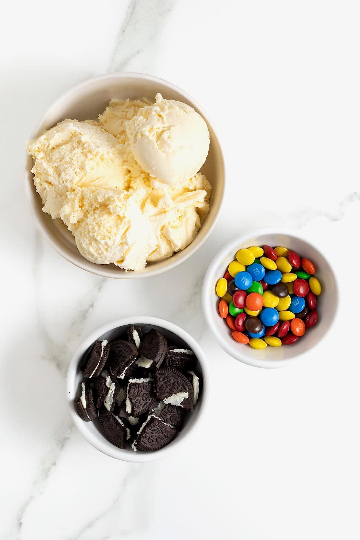 Ingredients to make a homemade Oreo and mini M&M blizzard in white dishes on a white marble counter.