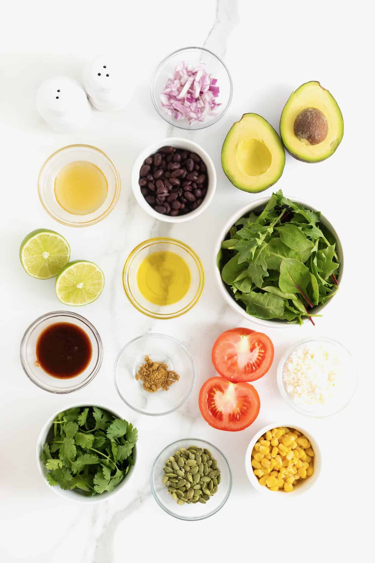 Ingredients to make a grilled avocado and tomato salad and a chipotle lime dressing in small glass dishes on a white marble counter.