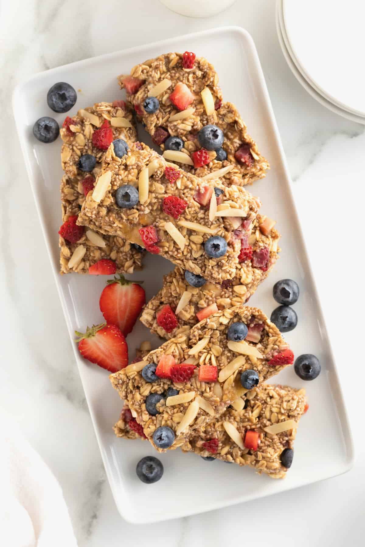A white rectangular serving platter stacked with overnight oats bars and blueberries and strawberry slices scattered around.
