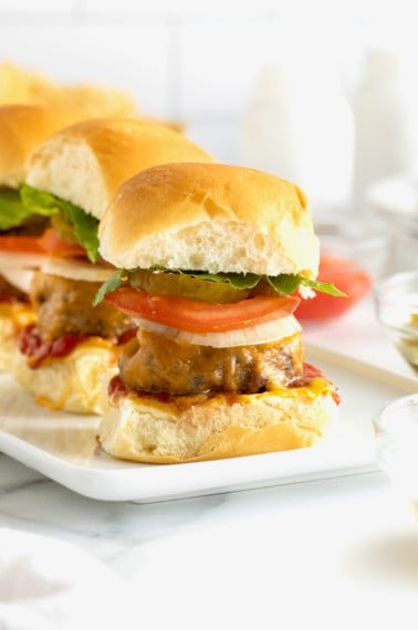Three mini cheeseburgers with pickle slices, tomato and lettuce on a white serving platter.