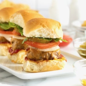 Three mini cheeseburgers with pickle slices, tomato and lettuce on a white serving platter.