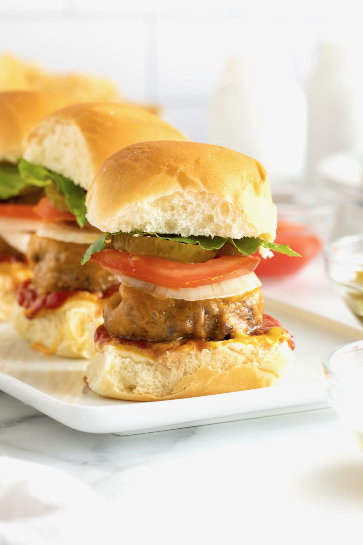 Three mini cheeseburgers with white onion, tomato,  pickles and lettuce on a white serving platter.