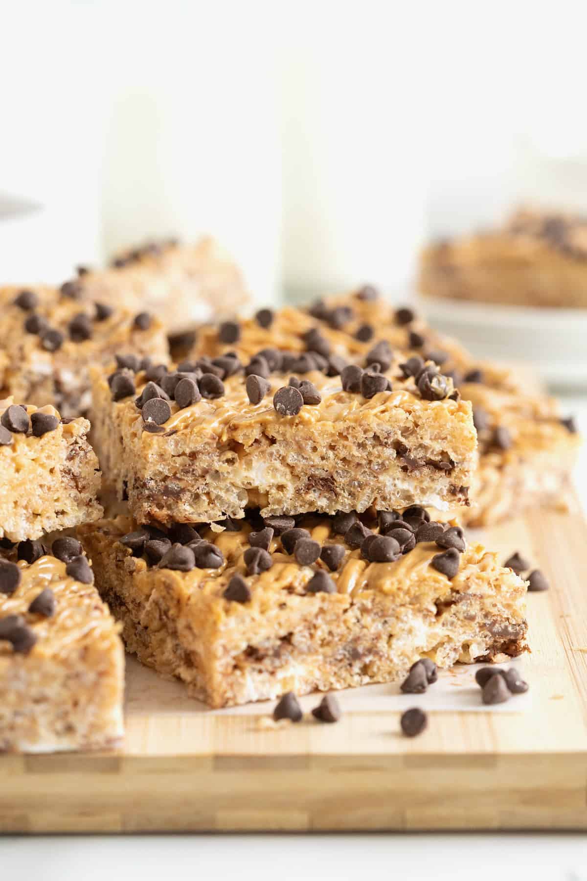 A pile of chocolate chip peanut butter Rice Krispies treats on a light wood cutting board. The top treat has a bite out of it.