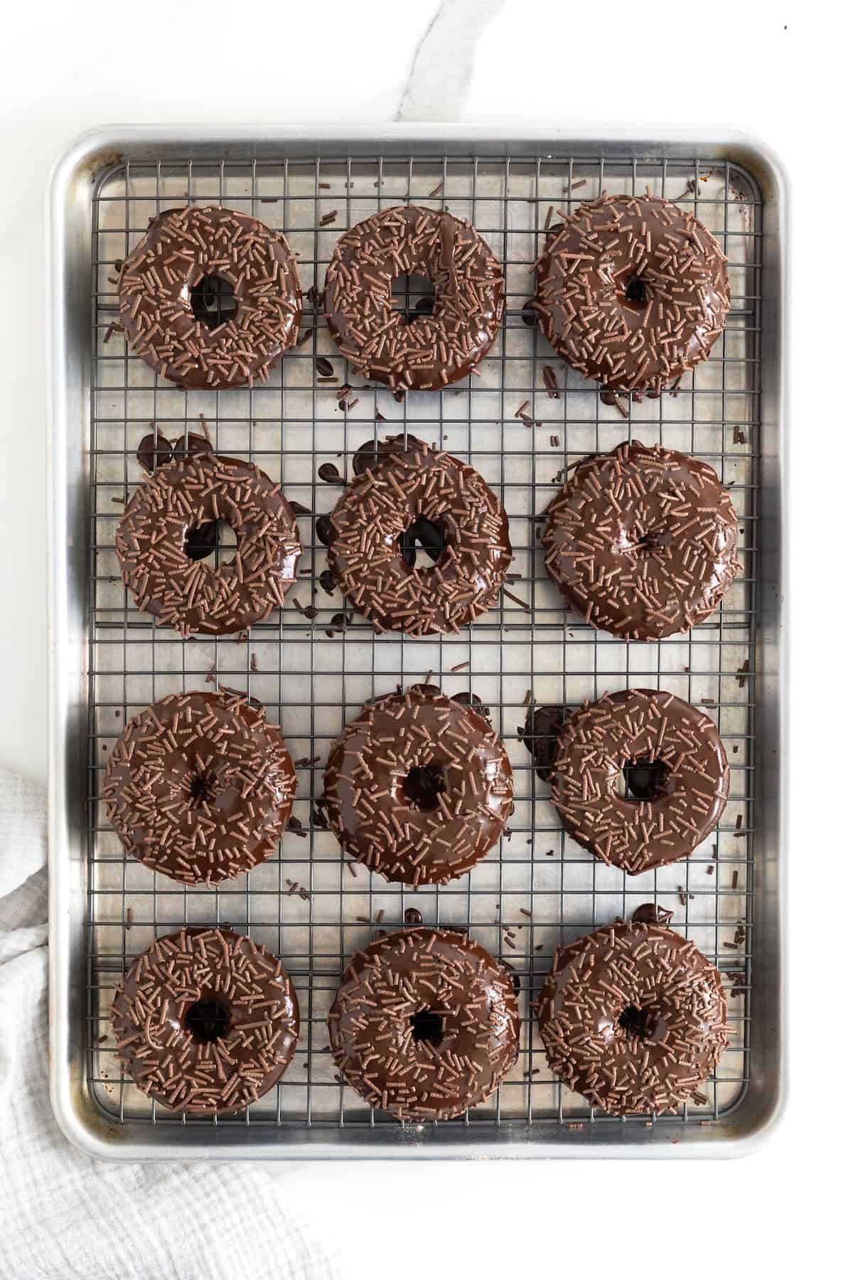 A dozen chocolate glazed chocolate donuts with chocolate sprinkles on a wire cooling rack.