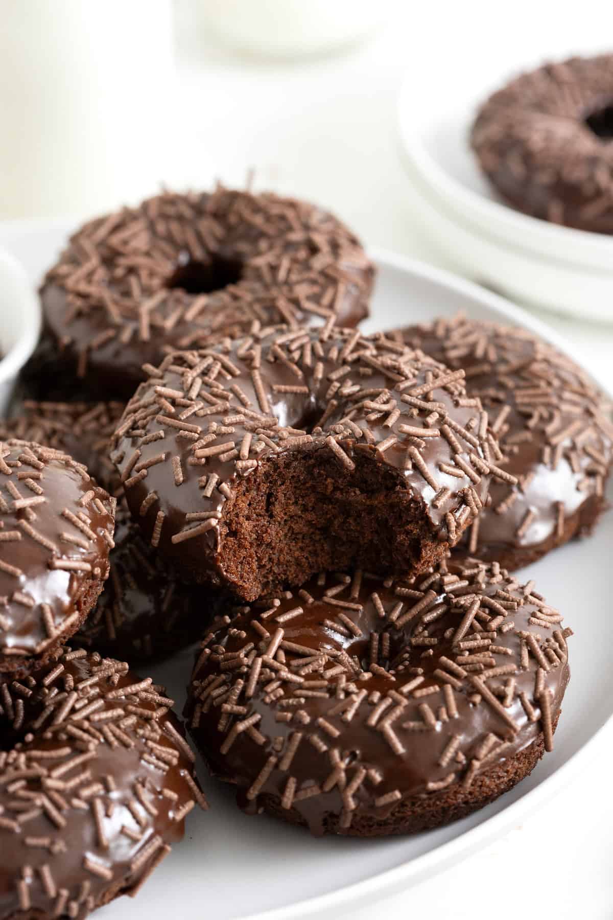 A pile of triple chocolate donuts on a white platter. The top donut has a bite out of it revealing a soft cake center.