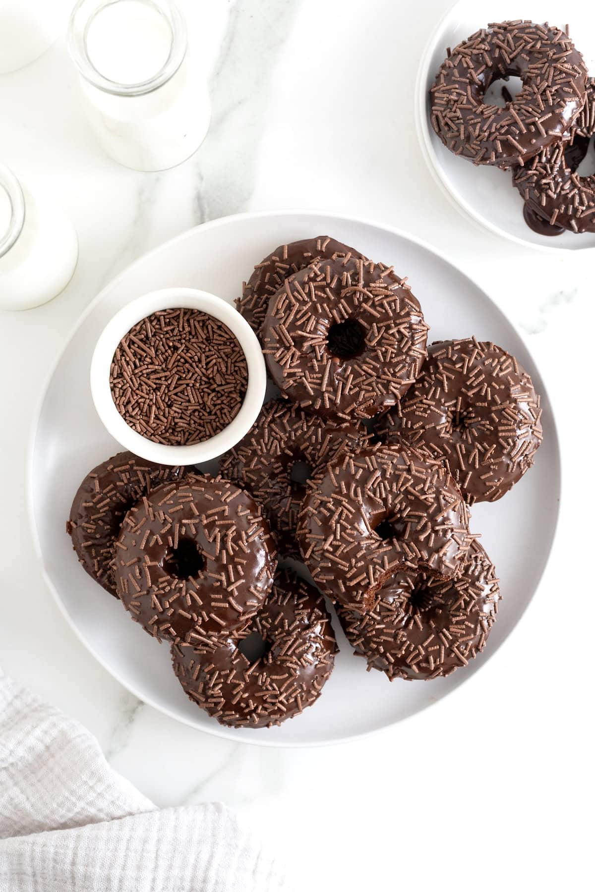 A pile of triple chocolate donuts on a white platter with a small bowl of chocolate sprinkles.