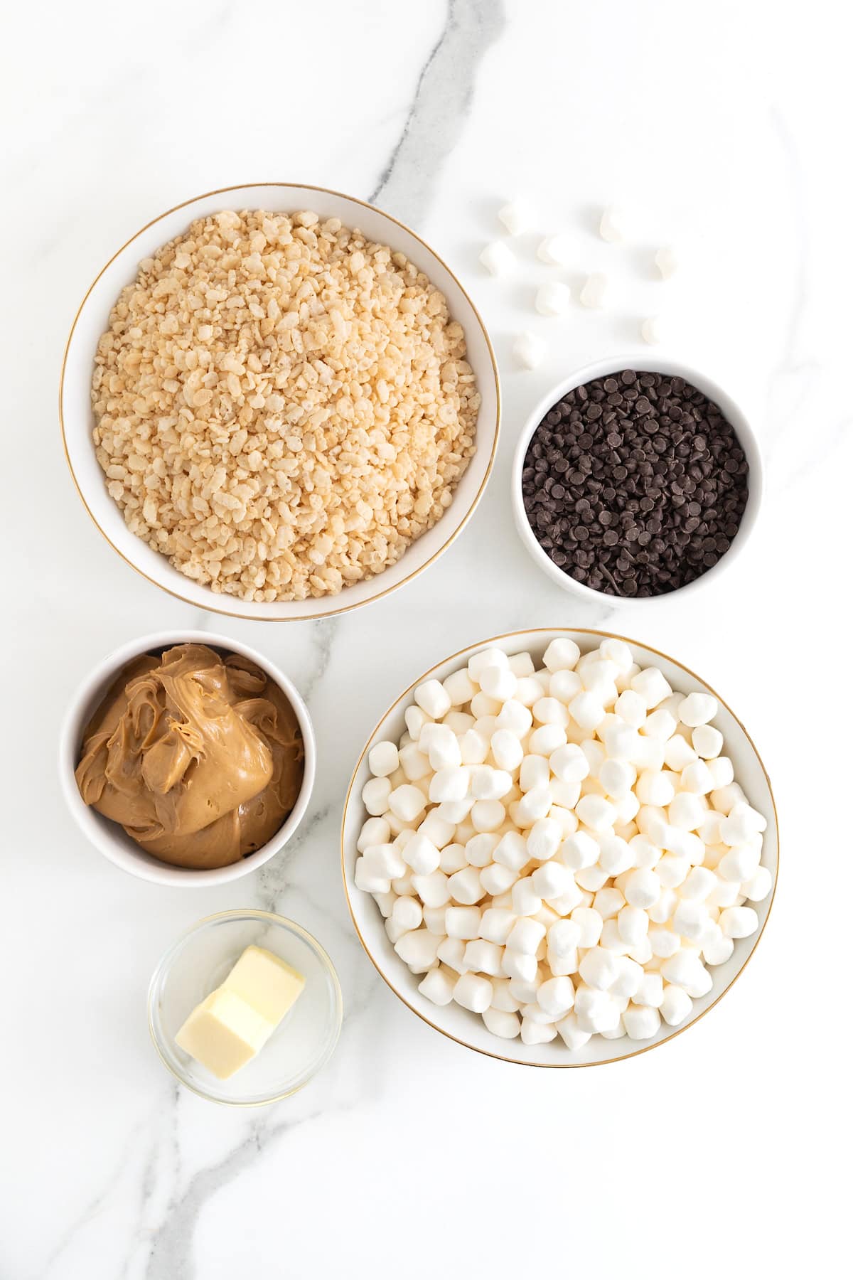 Ingredients to make peanut butter chocolate chip Rice Krispies treats in small white dishes on a white marble counter.