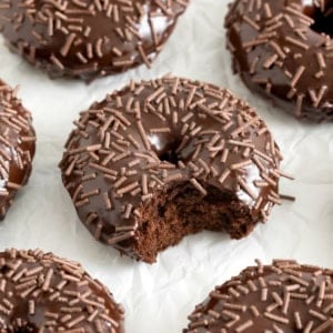 Seven chocolate donuts with chocolate icing and sprinkles on a piece of parchment paper. The center donut has a bite out of it.