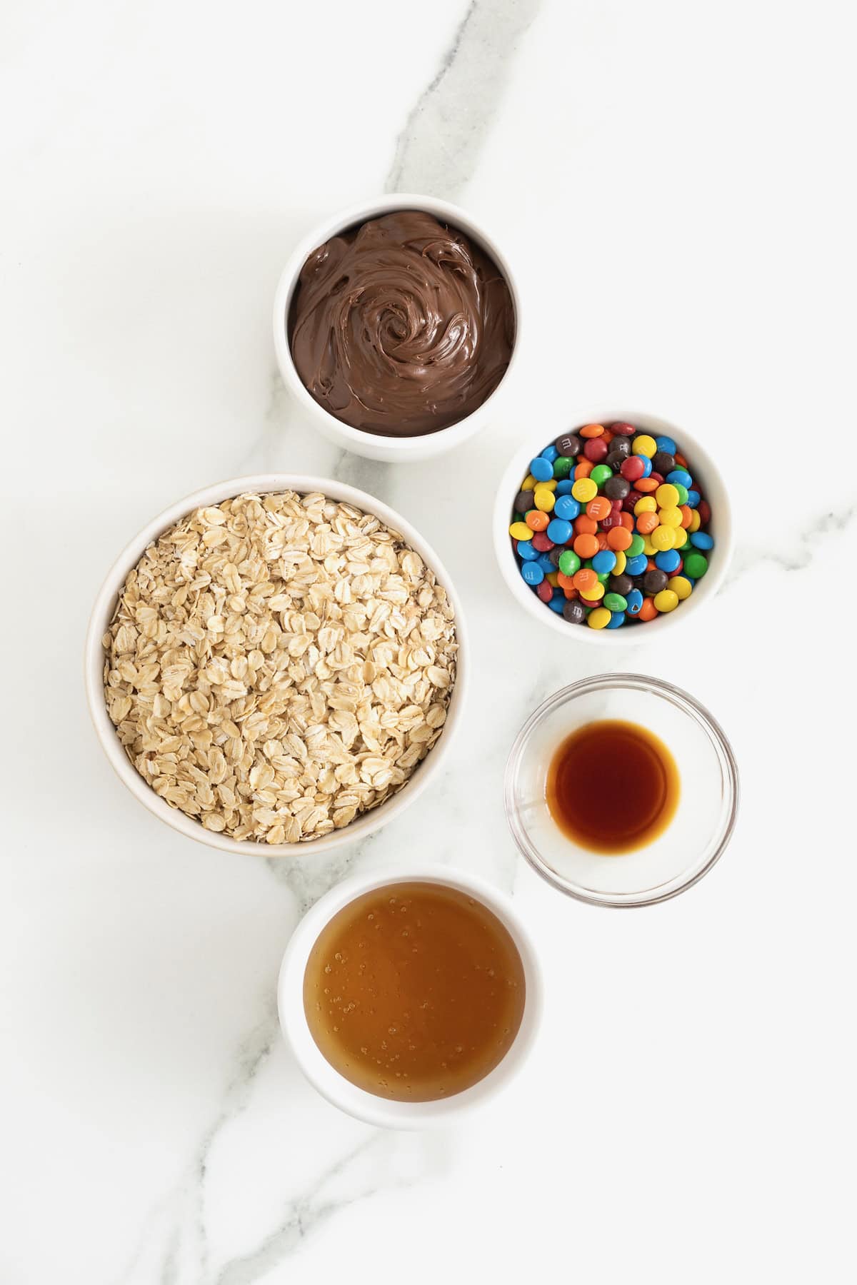 Ingredients to make Nutella M&M Oatmeal Balls in small glass dishes on a white marble counter.
