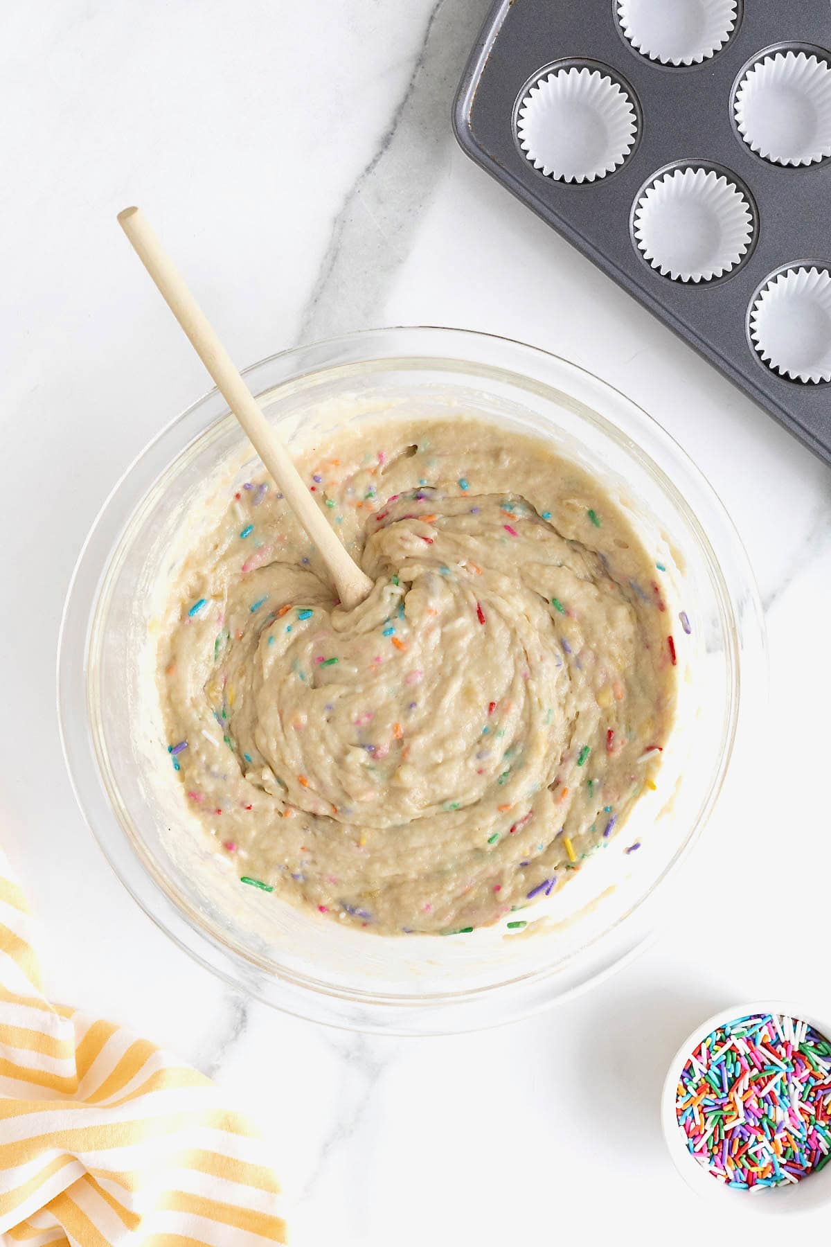 Rainbow sprinkles in muffin batter in a large clear glass mixing bowl.