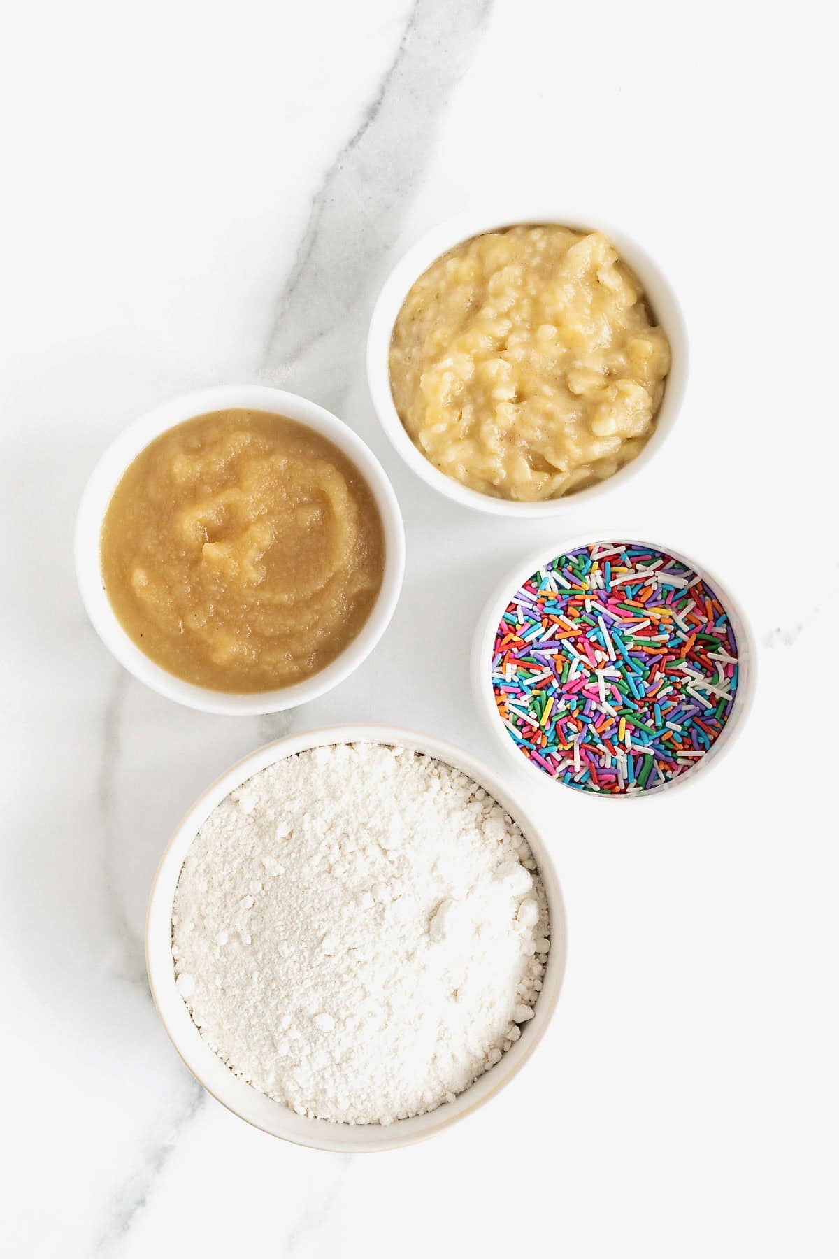Ingredients to make birthday breakfast mini muffins in small glass dishes on a white marble counter.