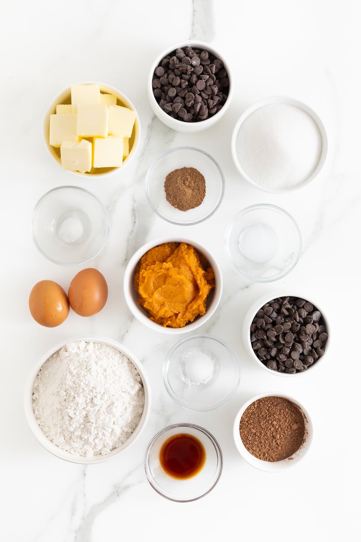 Ingredients to make triple chocolate pumpkin cookies in small glass dishes on a white marble counter.
