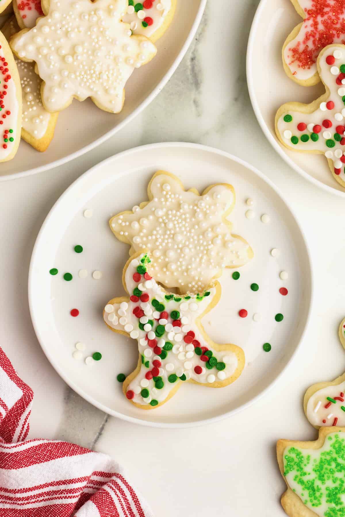 A white plate with two ice sugar cookies on it. One is a star with shite sprinkles and the other is a gingerbread man shape with red, white and green sprinkles. The gingerbread man has a bite out of it.